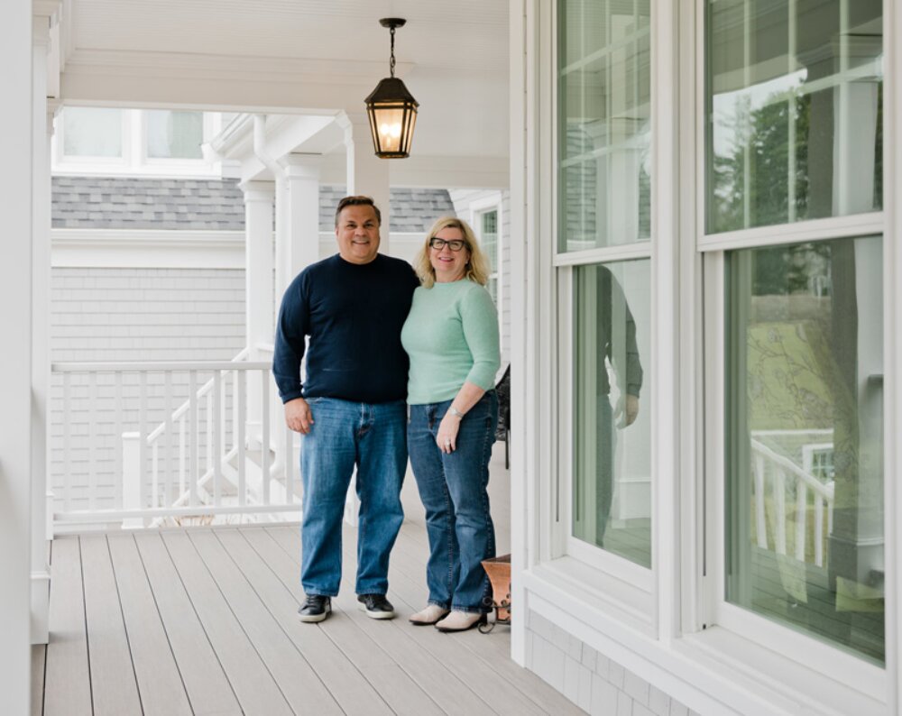 A photo of a smiling couple standing on the covered porch of a large home.
