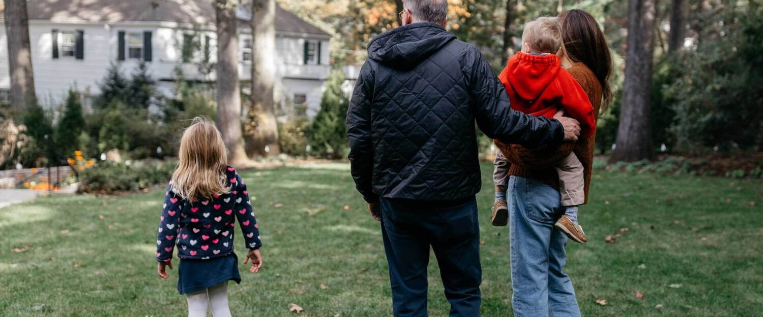 A photograph of a small family outside, facing toward their large white house.