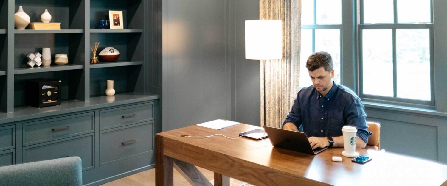 A photo of a man working at a laptop in a large, bright home office.
