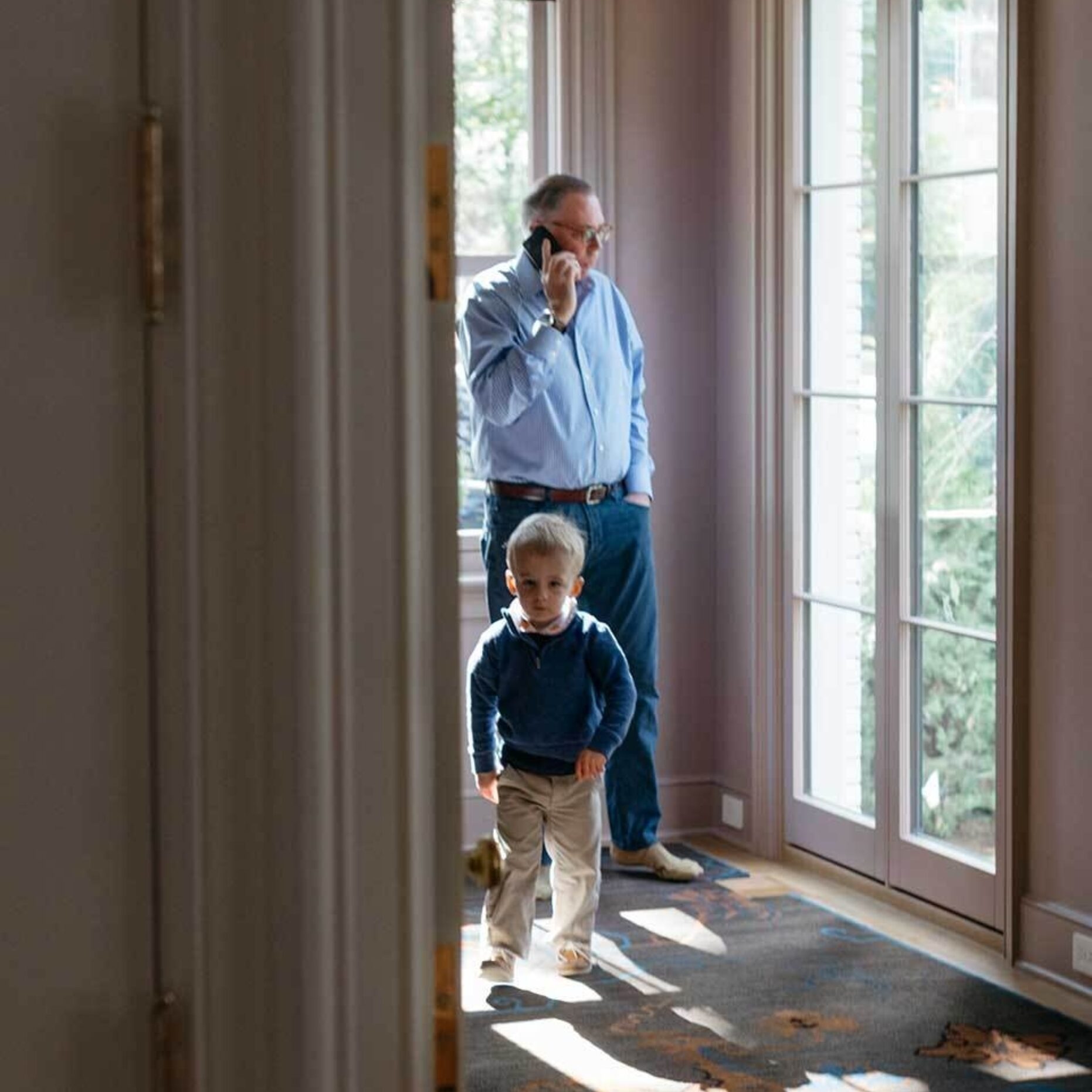 A man at home standing in front of a window, talking intently on a cellphone.
