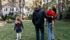 A photograph of a small family outside, facing toward their large white house.