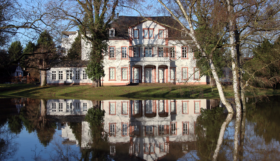A photo of the exterior of a large, luxury home with a flooded front yard.