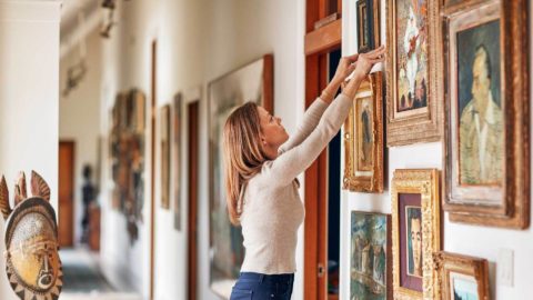 A photo of a woman adjusting a painting on the wall of a large home gallery.