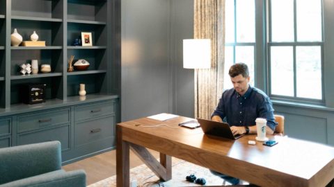 A photo of a man working at a laptop in a large, bright home office.