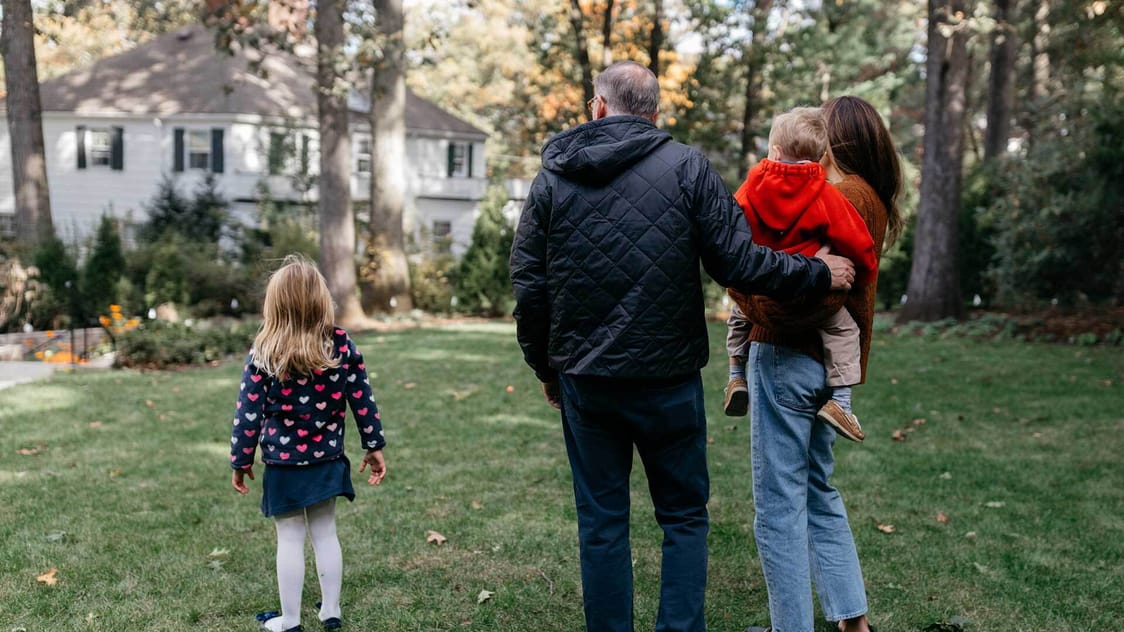 A photograph of a small family outside, facing toward their large white house.