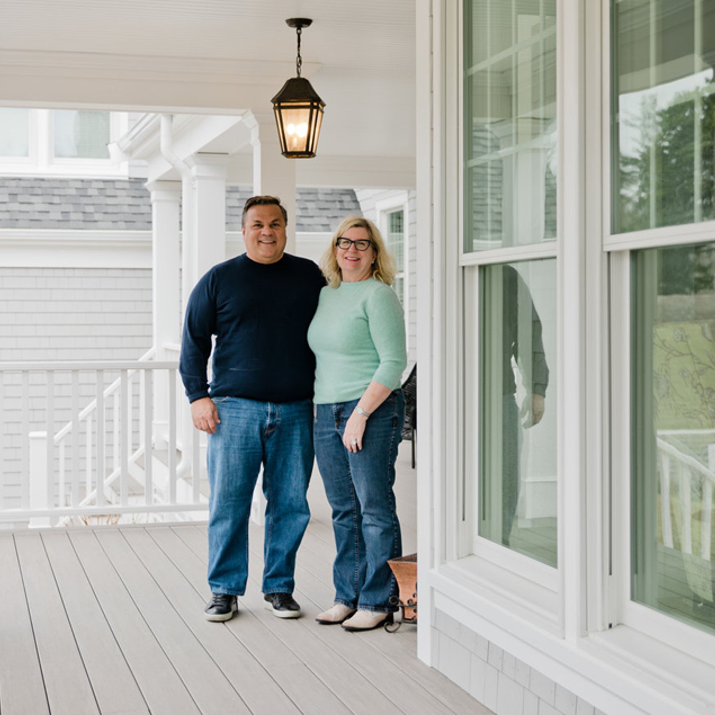 A photo of a smiling couple standing on the covered porch of a large home.