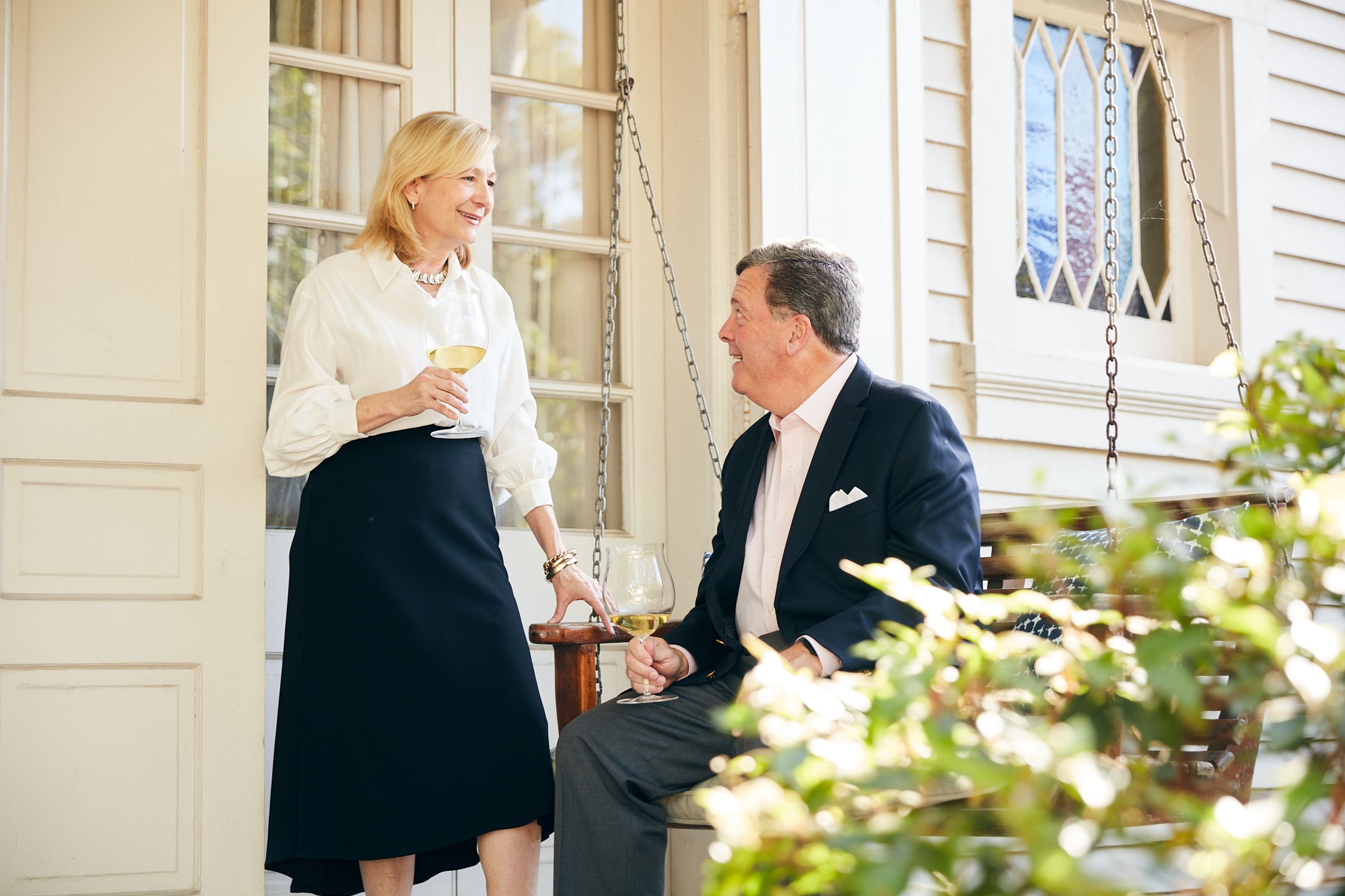 A couple looking and smiling at each other on the front porch of a white house with a wooden swing
